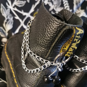 Op de afbeelding: Zwarte leren laarzen met zilveren kettingen en zwarte kralen. De laarzen hebben gele stiksels en een geel en zwart logo op de achterkant.