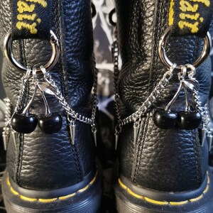 May include: Black leather boots with silver chains and cherry charms. The boots have yellow stitching and a thick black sole.