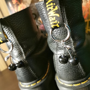 May include: Black leather boots with yellow stitching and a black cherry charm attached to a silver ring. The boots have a black strap with the text "Airwalk" in yellow.