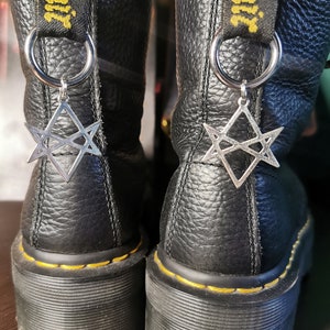 Könnte beinhalten: Ein Paar schwarze Lederstiefel mit gelben Nähten und einem silbernen Sternanhänger an jedem Stiefel. Die Stiefel haben eine dicke Sohle und einen schwarzen Lederriemen mit dem Text "Dr. Martens" darauf.