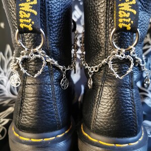 May include: A pair of black leather ankle boots featuring yellow AirWair heel loops and silver-toned metal chains. The chains are adorned with barbed wire heart charms and small pentagram pendants attached to the back of each boot.