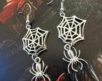 May include: A pair of silver spider web earrings with a dangling spider charm. The earrings are made of metal and have a delicate design.