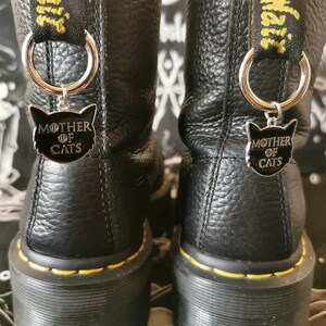 Könnte beinhalten: Ein Paar schwarze Leder-Plattformstiefel mit gelben Nähten und einem schwarzen und silbernen Anhänger mit der Aufschrift "Mutter der Katzen".