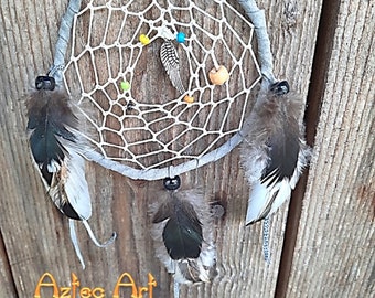 May include: A white dreamcatcher with grey and white feathers hanging from it. The dreamcatcher has a grey leather strap and is decorated with colourful beads. The dreamcatcher is hanging on a wooden surface. The text "Aztec Art" is visible in the bottom left corner of the image.