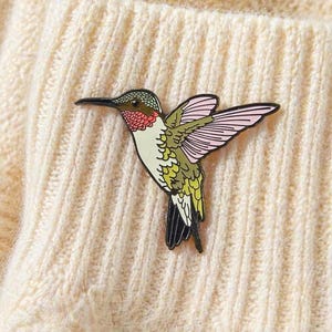 May include: A colorful hummingbird pin with a black outline. The pin features a green head, red throat, white chest, and green and pink wings. The hummingbird is set against a cream-colored, textured background.