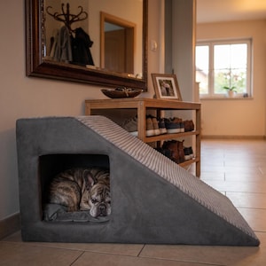 Puede incluir: Una rampa para mascotas de color gris oscuro con un hueco integrado que cuenta con un cojín interior suave. La rampa tiene una superficie texturizada y acanalada en la inclinación, y un perro atigrado descansa en el espacio cerrado.