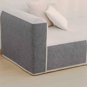 May include: A modern sectional sofa with a gray textured fabric on the arms and base, contrasted by a white seat and back. Two white pillows are placed on the sofa, creating a clean and minimalist aesthetic.