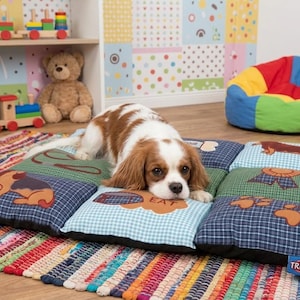 May include: A Cavalier King Charles Spaniel dog rests on a patchwork dog bed with various dog-themed designs. The bed is on a colorful striped rug. A wooden toy train and teddy bear are visible in the background.