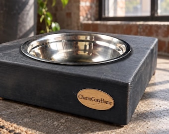 May include: A square, dark grey wooden pet bowl stand featuring a circular cutout holding a polished stainless steel bowl. An oval-shaped wooden plaque with the text CharmCozyHome is attached to the front side of the dark grey wooden base.