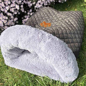 Puede incluir: Una cama gris para mascotas con un interior de felpa y un exterior acolchado. La cama tiene un parche marrón con una huella de pata. La cama está sobre hierba verde con flores moradas en el fondo.