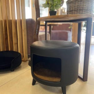 May include: A black pet bed with a rounded top and a cut-out opening. The bed has a brown cushion inside and sits on three short legs. A smaller black pet bed is visible in the background.
