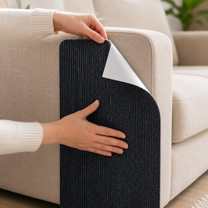 May include: A black, textured adhesive furniture protector being applied to the arm of a beige sofa. The protector has a peel-off backing and is designed to prevent scratches and wear.