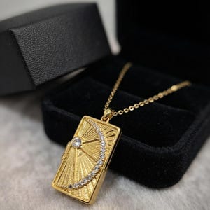 May include: A gold-toned rectangular pendant necklace featuring a sunburst design with a central clear stone and a crescent moon shape accented with small clear crystals. The pendant hangs from a delicate gold-toned chain.
