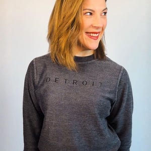 May include: A gray long-sleeved sweatshirt with the word "DETROIT" printed across the chest. The sweatshirt has a ribbed texture and is paired with blue jeans. The overall style is casual and comfortable.