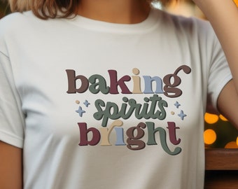 May include: A white t-shirt with the text "baking spirits bright" in colorful, stylized lettering. The shirt is worn by a person with brown hair and red lipstick, standing in front of a Christmas tree with lights.
