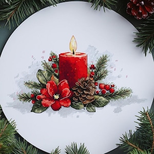 Può includere: Un disegno a punto croce di una candela rossa con una fiamma gialla, circondata da verde, bacche rosse e una pigna. Il disegno è su uno sfondo circolare bianco, incorniciato da rami di albero di Natale e ornamenti rossi.