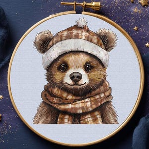 Puede incluir: Bordado de punto de cruz de un oso pardo con gorro y bufanda de invierno. El oso tiene una expresión amigable y está sobre un fondo azul claro dentro de un aro de bordado dorado. El diseño incluye el texto "Vihola CROSS STITCH".