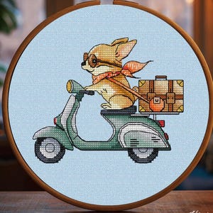 May include: Cross-stitch embroidery of a chihuahua dog riding a green scooter, wearing glasses and a scarf. The dog is carrying a suitcase. The embroidery is set in a wooden hoop against a light blue background.