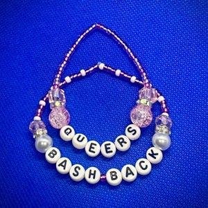 Könnte beinhalten: Eine rosa und weiße Perlenkette mit den Worten "Queers Bash Back" in schwarzen Buchstaben auf weißen Perlen.