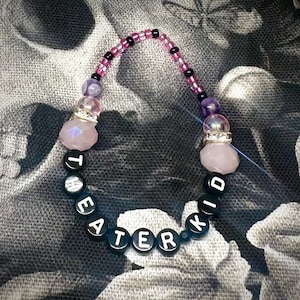 Op de afbeelding: Een kralenarmband met het woord "TEATER KID" gespeld met zwarte en witte letterkralen. De armband heeft roze en paarse kralen en roze gefacetteerde kristallen kralen. De achtergrond is een zwart-wit bloemenpatroon.