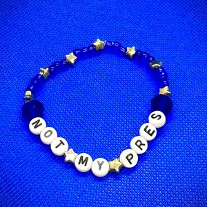 Könnte beinhalten: Ein blaues und weißes Perlenarmband mit goldenen Sternen und den Worten "NOT MY PRES" darauf.