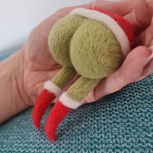 Peut inclure: Ornement de Noël en feutre fantaisiste représentant une figurine verte avec un chapeau et des jambes rouges. L'ornement a une bordure blanche sur le chapeau et les jambes, et des pieds rouges. La figurine est tenue dans une main.
