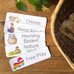 Pode incluir: Cartões retangulares brancos com ilustrações e texto, incluindo "Chores" e "Free Play". Os cartões estão sobre uma superfície de madeira. Uma cesta de vime e uma planta verde são visíveis no fundo. Perfeito para organização.