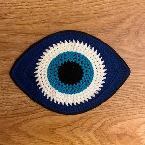 Könnte beinhalten: Ein handgefertigter, gehäkelter Untersetzer in Augenform. Der Untersetzer hat einen blauen Rand, einen weißen Ring, einen türkisfarbenen Ring und eine schwarze Mitte. Der Untersetzer liegt auf einer Holzoberfläche.