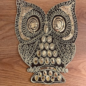 May include: A gold sequined owl applique with a brown felt backing. The owl has large, round eyes and a detailed design.