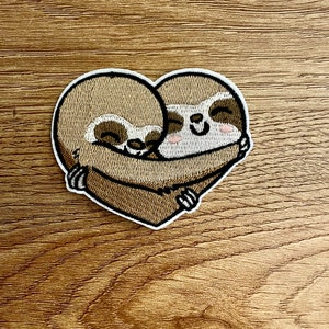 Pode incluir: Patch bordado com dois bichos-preguiça abraçando-se em forma de coração. Os bichos-preguiça são castanhos claros com detalhes em castanho escuro e rostos sorridentes. O patch tem uma borda branca e é projetado para aplicação em roupas ou acessórios.