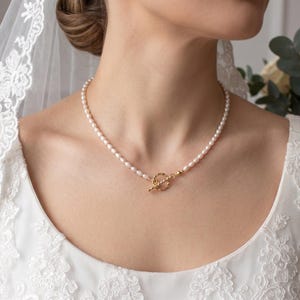 Peut inclure: Un collier de perles délicat avec un fermoir doré. Le collier est porté par une personne vêtue d'une robe et d'un voile blancs. Les perles sont petites et espacées uniformément, créant un look classique et élégant.