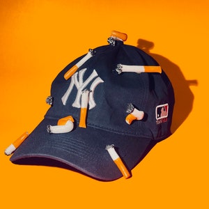 May include: A navy blue baseball cap with the New York Yankees logo in white. The cap is adorned with several cigarette-shaped embellishments, featuring white and orange details. The MLB logo is visible on the side.