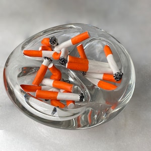 May include: A glass ashtray filled with orange and white painted cigarettes. The ashtray is clear and has a rounded shape.