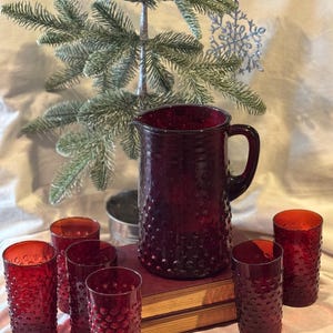 May include: A ruby red glass pitcher and matching drinking glasses with a textured, bubble-like pattern. The pitcher has a handle and is surrounded by six glasses. The glassware is displayed on a stack of books, with a small Christmas tree in the background.