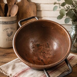 May include: A round, weathered copper bowl featuring two dark metal handles on opposite sides. The interior surface shows signs of oxidation and patina, with a deep reddish-brown metallic finish. The bowl rests on a textured, off-white fabric cloth.