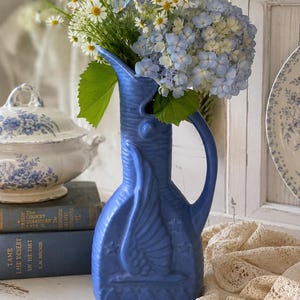 May include: A matte blue ceramic pitcher featuring a relief design of a bird in flight surrounded by small stars. The pitcher has a tall, slender neck, a curved handle, and is filled with a bouquet of white daisies and light blue hydrangeas.