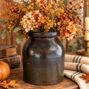 May include: A dark brown ceramic crock filled with an arrangement of dried autumn flowers, including orange and yellow hydrangeas, small berries, and green leaves. The cylindrical vessel has a smooth, glossy finish and a wide, thick rim.