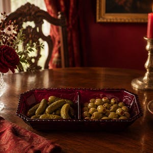 May include: A rectangular, deep red glass serving dish with a central divider, featuring a scalloped edge. The dish is filled with green pickles on one side and pimento-stuffed green olives on the other, showcasing a translucent, pressed glass texture.
