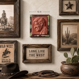May include: A square terracotta-colored wall plaque featuring a relief portrait of a person in profile, framed by a green and brown border. The plaque measures 6 inches in width and is displayed among other framed wall art and western decor.
