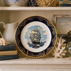 May include: A round ceramic decorative plate featuring a central illustration of a large sailing ship on blue water under a cloudy sky. The plate has a wide, dark navy blue border decorated with intricate gold filigree patterns and a gold rim.