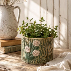 May include: A cylindrical ceramic planter with a textured green bark-like surface and three raised, pale pink flower appliques. The planter contains a small, leafy green plant with thin stems and numerous small, oval-shaped leaves.