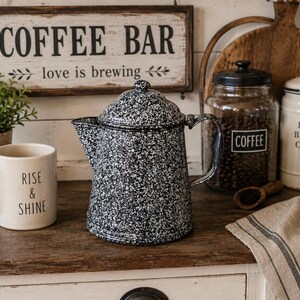 May include: A black and white speckled enamel coffee pot with a lid and handle, positioned next to a cream-colored ceramic mug with the text "RISE & SHINE" printed in black, alongside a glass jar labeled "COFFEE" and a white canister.