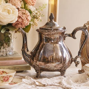 May include: A vintage silver teapot with ornate detailing, set on a lace tablecloth. The teapot is surrounded by a teacup and saucer, a small decorative dish, a framed picture, and a vase of flowers. The teapot is approximately 10 inches tall.