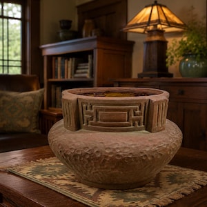 May include: A terracotta-colored ceramic bowl with a wide, rounded base and a textured, dimpled surface. The upper rim features a raised, geometric Greek key pattern integrated into the structure, creating a distinct, wide-mouthed opening.