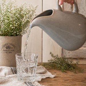 May include: A gray ceramic pitcher pouring water into a clear, patterned glass. The glass sits on a linen cloth with a gray stripe. A potted plant with white flowers and the text "FARMER'S MARKET FRESH & LOCAL No. 1892" is in the background.