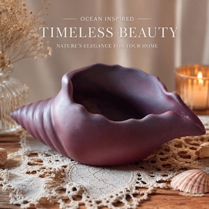 May include: A matte dark purple ceramic bowl shaped like a conch shell. The text above reads OCEAN INSPIRED TIMELESS BEAUTY NATURE'S ELEGANCE FOR YOUR HOME. The bowl has a smooth, curved surface with a wide opening and a tapered, spiraled end.