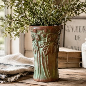 May include: A ceramic vase with a flared rim and a tapered base, featuring a raised relief pattern of leaves and stems. The surface has a weathered finish with blended tones of muted green and earthy brown, creating a textured appearance.