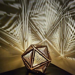 Może przedstawiać: Drewniana lampa o geometrycznym kształcie z włączonym światłem, rzucającym skomplikowane cienie na ścianę za nią.