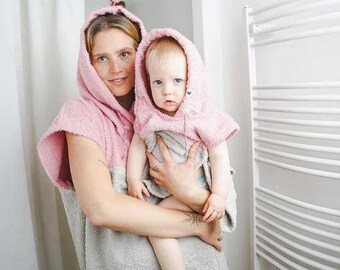 Könnte beinhalten: Eine Frau hält ein Baby, beide tragen passende rosa Kapuzenhandtücher. Die Handtücher sind an der Kapuze und den Schultern rosa, der Rest ist grau. Der Hintergrund ist ein Badezimmer mit weißem Vorhang und Handtuchhalter.