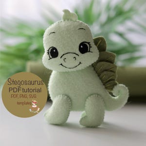May include: A light green felt stegosaurus dinosaur plush toy with embroidered details. The dinosaur has large black eyes, a smiling mouth, and a row of green plates along its back. The text "Stegosaurus PDF tutorial" is on a circular label.